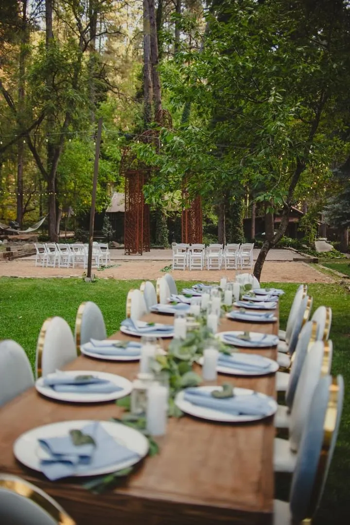 Long wooden farm table set with blue linens under string lights in a forest ceremony space
